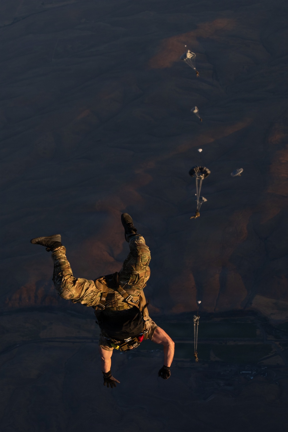 A military paratrooper in camouflage uniform is captured mid-freefall immediately after exiting an aircraft, photographed from above looking down toward arid, mountainous terrain far below. Several other paratroopers with partially deployed parachutes are visible at various altitudes in the background. The image captures a military static-line or HALO/HAHO jump operation over desert terrain.