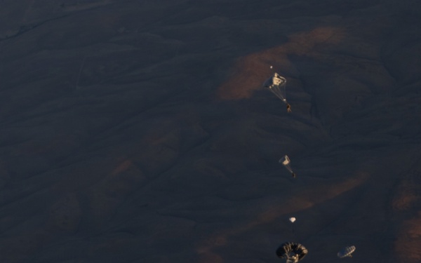 A military paratrooper in camouflage uniform is captured mid-freefall immediately after exiting an aircraft, photographed from above looking down toward arid, mountainous terrain far below. Several other paratroopers with partially deployed parachutes are visible at various altitudes in the background. The image captures a military static-line or HALO/HAHO jump operation over desert terrain.