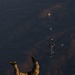 A military paratrooper in camouflage uniform is captured mid-freefall immediately after exiting an aircraft, photographed from above looking down toward arid, mountainous terrain far below. Several other paratroopers with partially deployed parachutes are visible at various altitudes in the background. The image captures a military static-line or HALO/HAHO jump operation over desert terrain.
