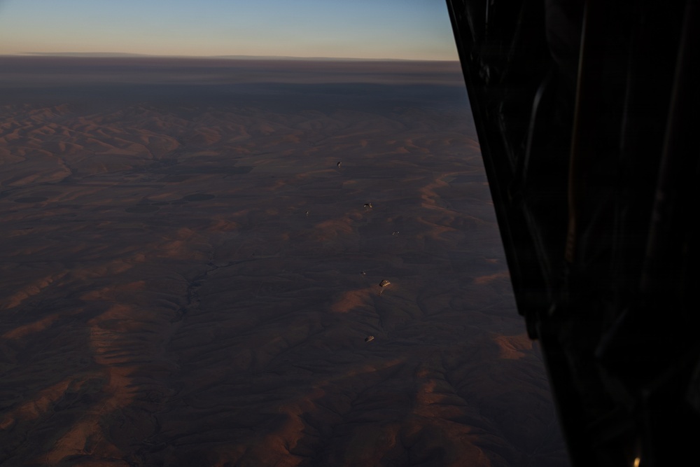 An aerial photograph taken from inside an aircraft looking out over a vast arid desert landscape at low light conditions, likely dawn or dusk. Several parachutes or paragliders are visible at various altitudes between the aircraft and the ground below. The terrain appears to be a dry, mountainous or hilly desert region with eroded ridgelines.