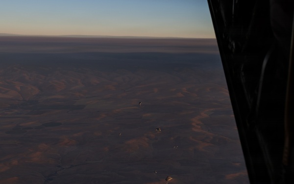 An aerial photograph taken from inside an aircraft looking out over a vast arid desert landscape at low light conditions, likely dawn or dusk. Several parachutes or paragliders are visible at various altitudes between the aircraft and the ground below. The terrain appears to be a dry, mountainous or hilly desert region with eroded ridgelines.