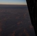 An aerial photograph taken from inside an aircraft looking out over a vast arid desert landscape at low light conditions, likely dawn or dusk. Several parachutes or paragliders are visible at various altitudes between the aircraft and the ground below. The terrain appears to be a dry, mountainous or hilly desert region with eroded ridgelines.