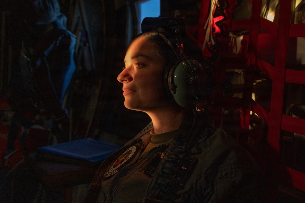 A military service member is seated inside what appears to be a military aircraft, wearing aviation headphones and a flight suit with visible patches. The subject is illuminated by warm directional lighting against a dark background, creating a dramatic portrait-style composition. The individual appears relaxed, looking slightly upward with a subtle expression.