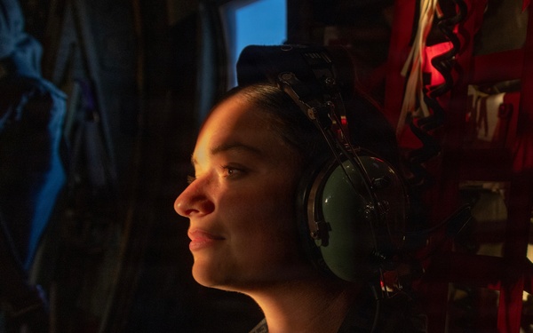 A military service member is seated inside what appears to be a military aircraft, wearing aviation headphones and a flight suit with visible patches. The subject is illuminated by warm directional lighting against a dark background, creating a dramatic portrait-style composition. The individual appears relaxed, looking slightly upward with a subtle expression.