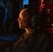A military service member is seated inside what appears to be a military aircraft, wearing aviation headphones and a flight suit with visible patches. The subject is illuminated by warm directional lighting against a dark background, creating a dramatic portrait-style composition. The individual appears relaxed, looking slightly upward with a subtle expression.
