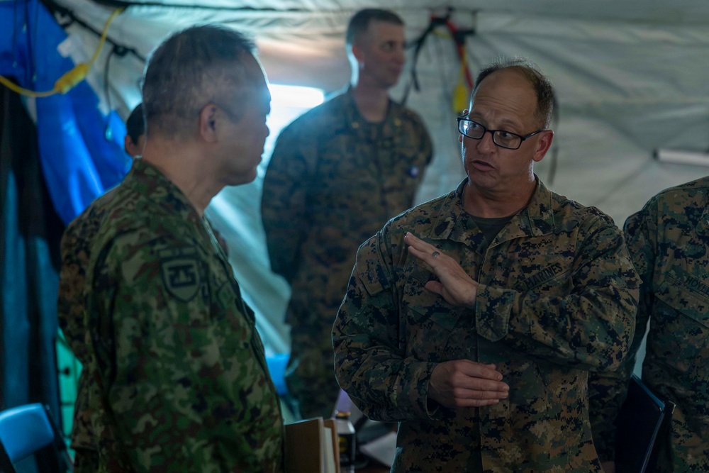 The image shows military personnel from at least two different armed forces engaged in conversation inside what appears to be a military field tent or command post. A U.S. Marine (identified by 'MARINES' tape on uniform and MARPAT digital camouflage) wearing glasses is speaking with a soldier in a different camouflage pattern, likely Japanese Ground Self-Defense Force based on the uniform style and patch. A third service member is visible in the background observing the discussion.