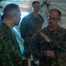 The image shows military personnel from at least two different armed forces engaged in conversation inside what appears to be a military field tent or command post. A U.S. Marine (identified by 'MARINES' tape on uniform and MARPAT digital camouflage) wearing glasses is speaking with a soldier in a different camouflage pattern, likely Japanese Ground Self-Defense Force based on the uniform style and patch. A third service member is visible in the background observing the discussion.