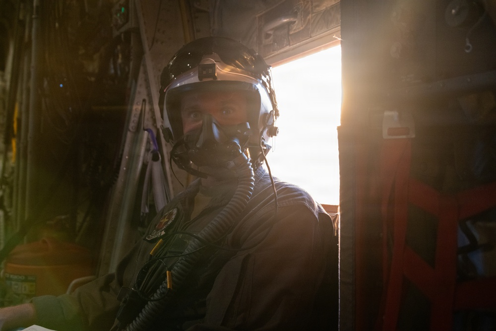 A military aviator or aircrew member is photographed inside an aircraft cabin, wearing full flight gear including a helmet, oxygen mask, and flight suit. Strong backlighting from an open door or window creates a dramatic lens flare effect. The subject appears to be looking directly at the camera while seated or positioned near the aircraft's interior.