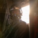 A military aviator or aircrew member is photographed inside an aircraft cabin, wearing full flight gear including a helmet, oxygen mask, and flight suit. Strong backlighting from an open door or window creates a dramatic lens flare effect. The subject appears to be looking directly at the camera while seated or positioned near the aircraft's interior.