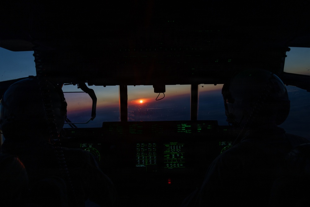 A cockpit view from a military or large transport aircraft during twilight, with two crew members silhouetted against a vivid sunset on the horizon. The instrument panel glows green with multiple illuminated displays and controls. The scene captures a low-light operational flight environment at dusk or dawn.