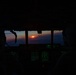 A cockpit view from a military or large transport aircraft during twilight, with two crew members silhouetted against a vivid sunset on the horizon. The instrument panel glows green with multiple illuminated displays and controls. The scene captures a low-light operational flight environment at dusk or dawn.