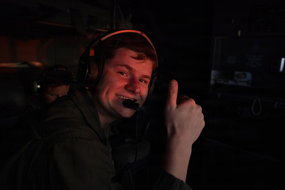 A young male is photographed in a dark environment wearing a gaming or communications headset with an orange accent band and a boom microphone. He is smiling and giving a thumbs-up gesture toward the camera. The scene appears to be a gaming setup or broadcast/production environment based on the visible monitor in the background.
