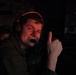 A young male is photographed in a dark environment wearing a gaming or communications headset with an orange accent band and a boom microphone. He is smiling and giving a thumbs-up gesture toward the camera. The scene appears to be a gaming setup or broadcast/production environment based on the visible monitor in the background.