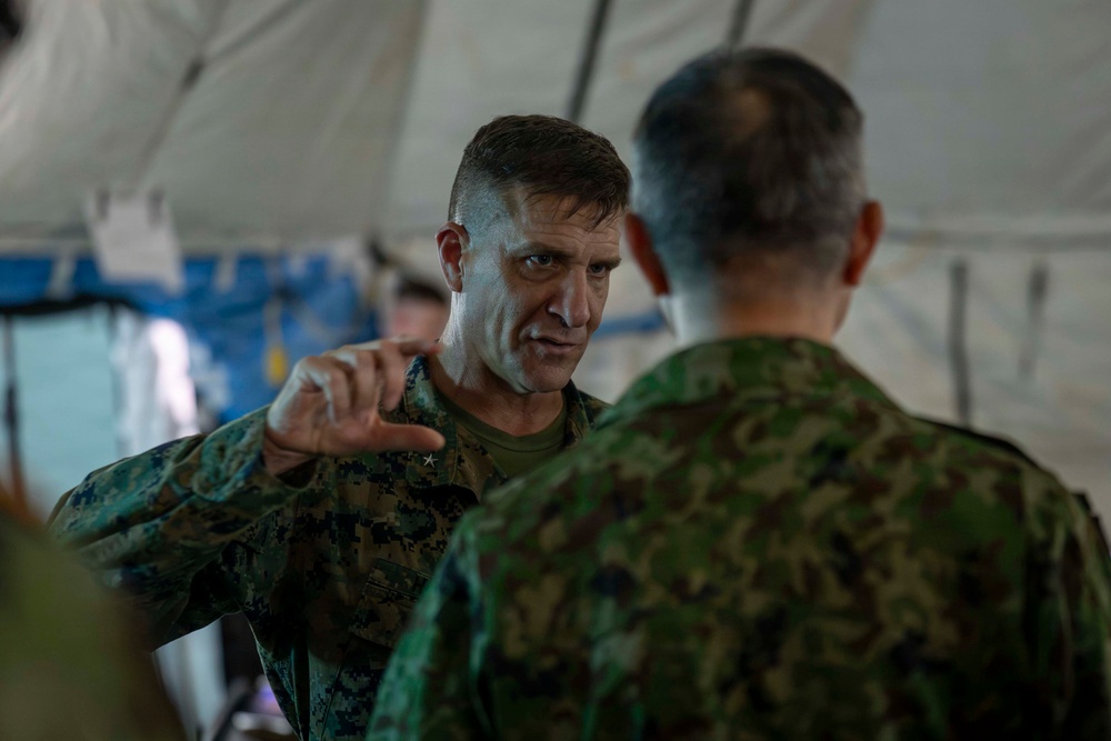 Two military personnel in camouflage uniforms are engaged in a discussion inside what appears to be a military tent or field structure. The individual facing the camera, wearing MARPAT (US Marine Corps digital camouflage) with a visible star insignia indicating general officer rank, is gesturing with his hand while speaking. The other individual, facing away, appears to be wearing a different camouflage pattern consistent with a foreign military uniform.