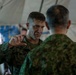 Two military personnel in camouflage uniforms are engaged in a discussion inside what appears to be a military tent or field structure. The individual facing the camera, wearing MARPAT (US Marine Corps digital camouflage) with a visible star insignia indicating general officer rank, is gesturing with his hand while speaking. The other individual, facing away, appears to be wearing a different camouflage pattern consistent with a foreign military uniform.