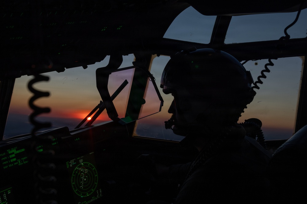 A military pilot is silhouetted in the cockpit of a large aircraft during sunset or sunrise. The cockpit features green-illuminated avionics displays and instrumentation, including a visible radar scope. The dramatic low-light conditions create a high-contrast scene with warm orange and red tones visible through the windscreen.