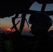 A military pilot is silhouetted in the cockpit of a large aircraft during sunset or sunrise. The cockpit features green-illuminated avionics displays and instrumentation, including a visible radar scope. The dramatic low-light conditions create a high-contrast scene with warm orange and red tones visible through the windscreen.