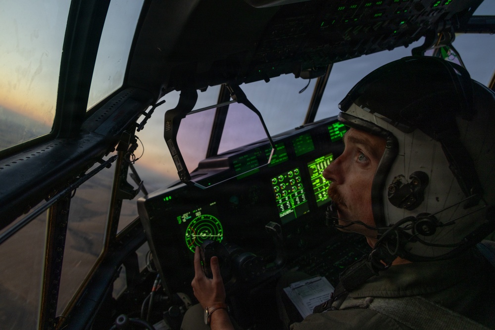 A military pilot is seated in the cockpit of a large aircraft during twilight or dawn conditions. The cockpit is illuminated by green-glowing instrument panels and displays, including what appears to be a radar screen. The pilot is wearing a flight helmet with oxygen mask equipment and appears to be monitoring instruments while in flight.