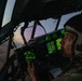 A military pilot is seated in the cockpit of a large aircraft during twilight or dawn conditions. The cockpit is illuminated by green-glowing instrument panels and displays, including what appears to be a radar screen. The pilot is wearing a flight helmet with oxygen mask equipment and appears to be monitoring instruments while in flight.