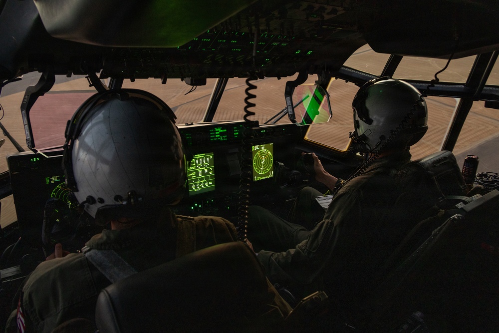 Two military aircrew members are seated in the cockpit of a large military aircraft, likely a C-130 Hercules or similar transport aircraft, operating during low-light or dusk conditions. The cockpit is illuminated primarily by green-glowing avionics displays and instrument panels. The exterior environment visible through the windscreen appears to be an arid, desert-like terrain.