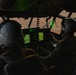Two military aircrew members are seated in the cockpit of a large military aircraft, likely a C-130 Hercules or similar transport aircraft, operating during low-light or dusk conditions. The cockpit is illuminated primarily by green-glowing avionics displays and instrument panels. The exterior environment visible through the windscreen appears to be an arid, desert-like terrain.