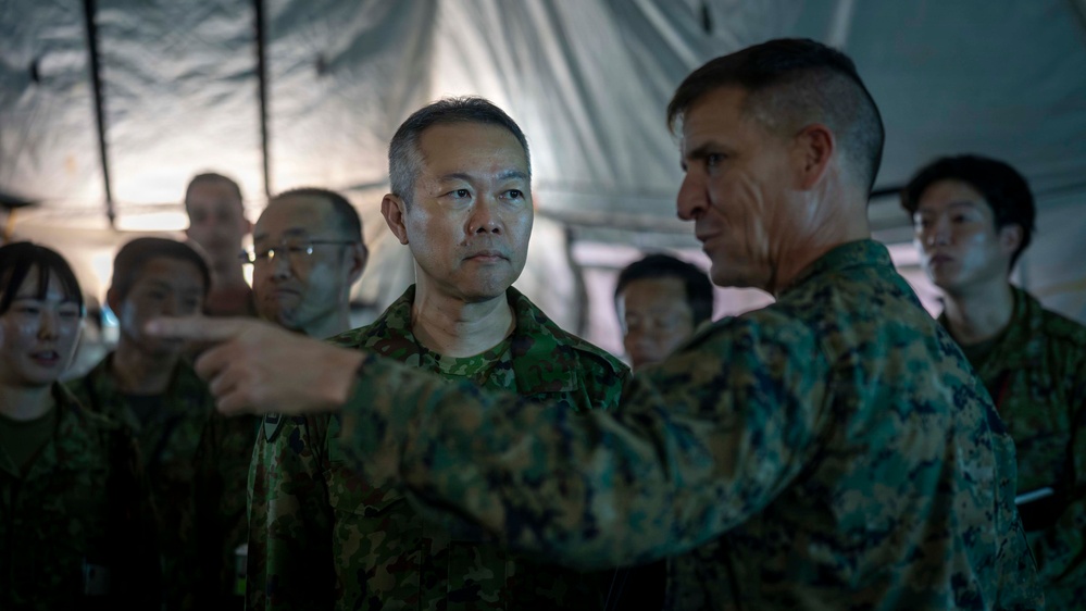 Two military officers in different camouflage uniforms appear to be engaged in a briefing or discussion inside a military tent. One officer, wearing what appears to be Japanese Ground Self-Defense Force camouflage, listens attentively while the other, in what appears to be US Marine Corps MARPAT camouflage, gestures as if explaining or pointing something out. Several other military personnel are visible in the background.