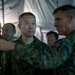 Two military officers in different camouflage uniforms appear to be engaged in a briefing or discussion inside a military tent. One officer, wearing what appears to be Japanese Ground Self-Defense Force camouflage, listens attentively while the other, in what appears to be US Marine Corps MARPAT camouflage, gestures as if explaining or pointing something out. Several other military personnel are visible in the background.