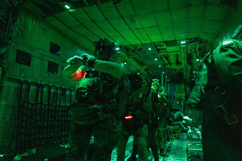 Military personnel in full tactical gear are positioned inside the cargo hold of a large military transport aircraft, illuminated by green and red interior lighting. The soldiers are equipped with night vision devices, tactical vests, and appear to be preparing for or staging a jump or insertion operation. The green lighting is consistent with pre-jump protocols used to preserve night vision adaptation.