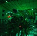 Military personnel in full tactical gear are positioned inside the cargo hold of a large military transport aircraft, illuminated by green and red interior lighting. The soldiers are equipped with night vision devices, tactical vests, and appear to be preparing for or staging a jump or insertion operation. The green lighting is consistent with pre-jump protocols used to preserve night vision adaptation.