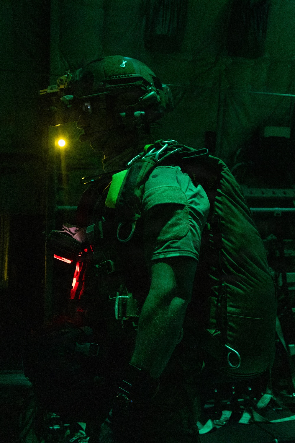A heavily equipped military or special operations personnel is photographed in a low-light environment illuminated by green and red ambient lighting. The individual is wearing tactical gear including a ballistic helmet with night vision device mounts, a large rucksack, and various harness attachments. The setting appears to be the interior of a military aircraft or vehicle.