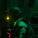 A heavily equipped military or special operations personnel is photographed in a low-light environment illuminated by green and red ambient lighting. The individual is wearing tactical gear including a ballistic helmet with night vision device mounts, a large rucksack, and various harness attachments. The setting appears to be the interior of a military aircraft or vehicle.