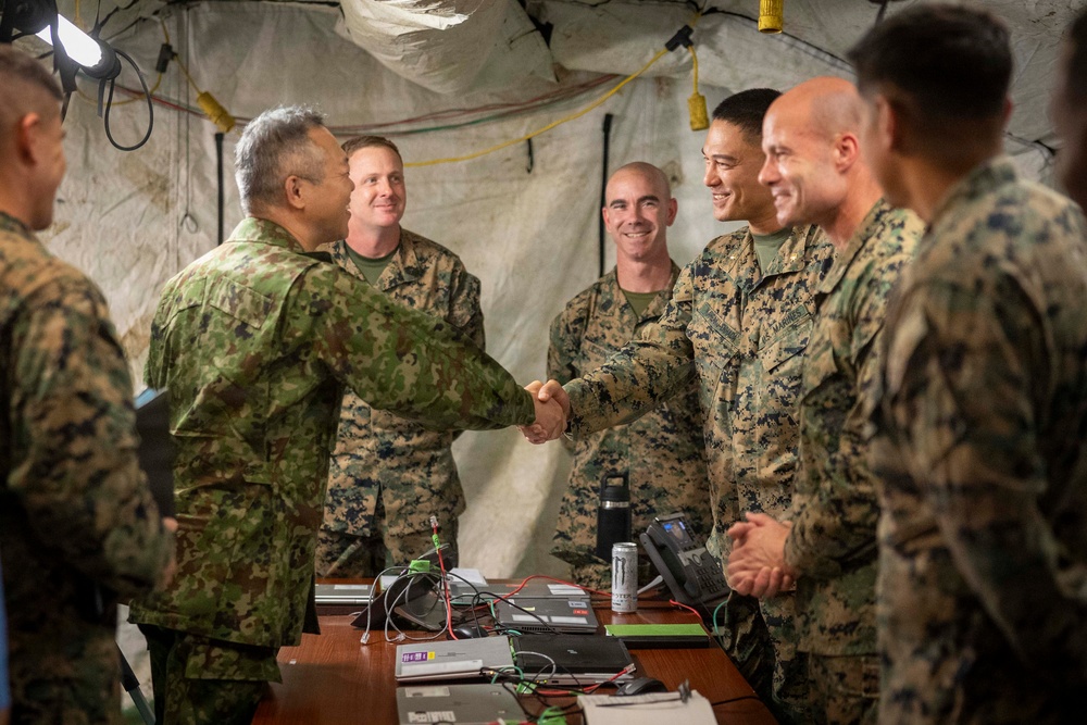 Military personnel from what appear to be two different armed forces are gathered inside a field command tent or temporary operations center. A handshake is taking place between a soldier in a distinctive camouflage pattern (consistent with Japan Ground Self-Defense Force) and a U.S. Marine in MARPAT digital camouflage. The atmosphere appears cordial and professional, suggesting a joint exercise or bilateral military engagement.