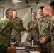 Military personnel from what appear to be two different armed forces are gathered inside a field command tent or temporary operations center. A handshake is taking place between a soldier in a distinctive camouflage pattern (consistent with Japan Ground Self-Defense Force) and a U.S. Marine in MARPAT digital camouflage. The atmosphere appears cordial and professional, suggesting a joint exercise or bilateral military engagement.