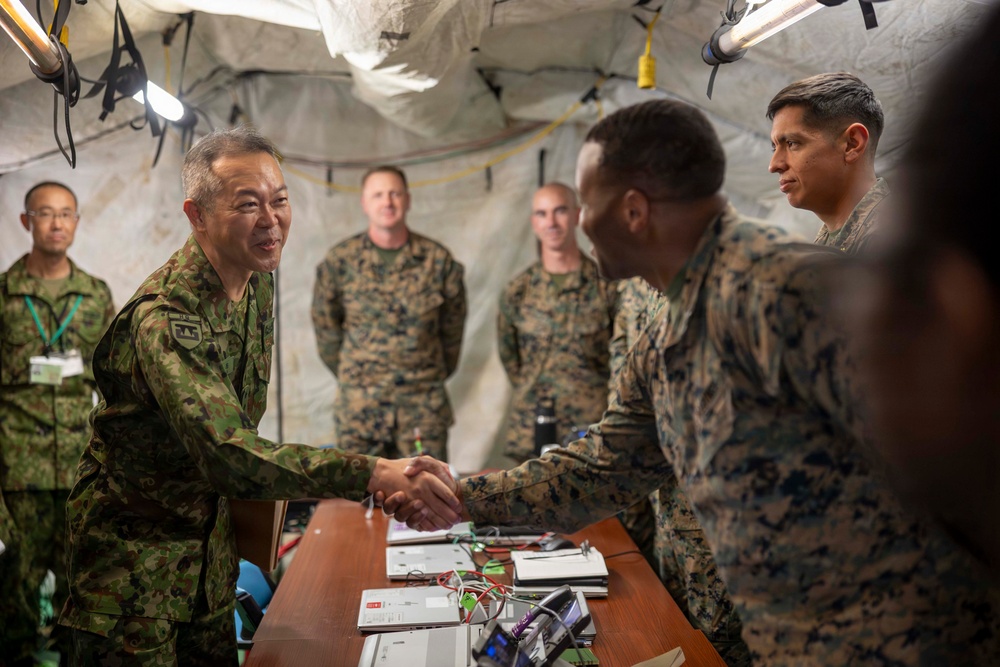 Military personnel from what appear to be two different nations are meeting inside a field tent or temporary command structure. A senior officer in a distinctive camouflage pattern consistent with the Japan Ground Self-Defense Force is shaking hands with a U.S. Marine in MARPAT camouflage across a table with electronic equipment. The setting suggests a joint military exercise or official liaison visit.