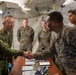 Military personnel from what appear to be two different nations are meeting inside a field tent or temporary command structure. A senior officer in a distinctive camouflage pattern consistent with the Japan Ground Self-Defense Force is shaking hands with a U.S. Marine in MARPAT camouflage across a table with electronic equipment. The setting suggests a joint military exercise or official liaison visit.