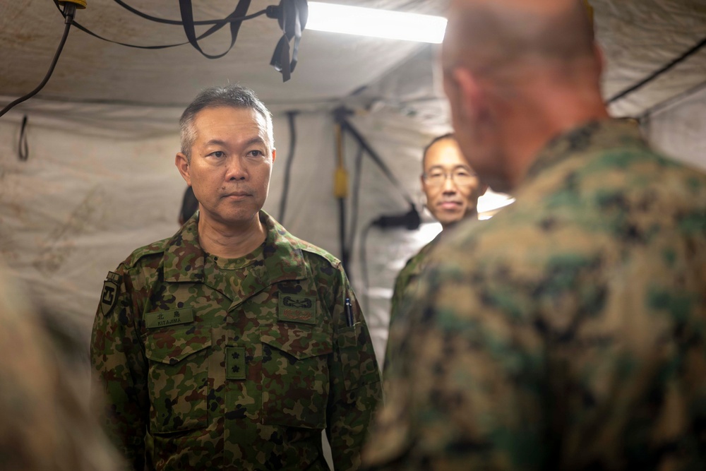 The image shows military personnel inside a tent or temporary command structure, appearing to be engaged in a briefing or discussion. At least two individuals are wearing Japanese Ground Self-Defense Force (JGSDF) camouflage uniforms, while a third person in the foreground (seen from behind) appears to be wearing US Marine Corps MARPAT camouflage. The setting suggests a joint military exercise or coordination meeting.