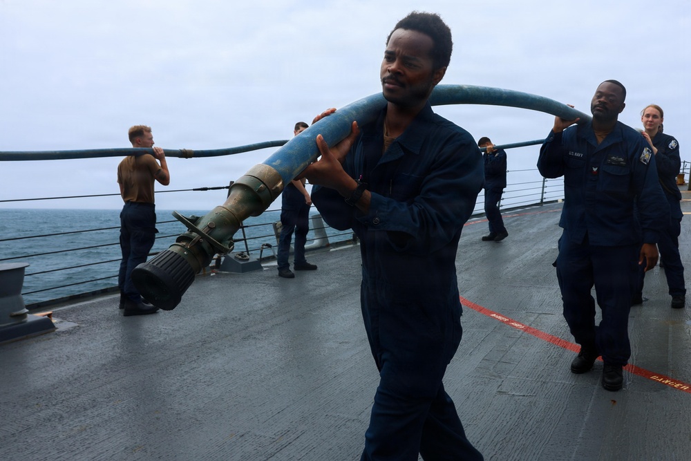 Naval personnel in dark blue coveralls are working on the deck of a military vessel at sea, carrying and handling a large blue hose with a metal nozzle/coupling attachment. The scene appears to be a training exercise or operational drill conducted on an overcast day. Multiple crew members are visible on the ship's deck, with the ocean visible in the background.
