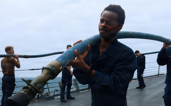 Naval personnel in dark blue coveralls are working on the deck of a military vessel at sea, carrying and handling a large blue hose with a metal nozzle/coupling attachment. The scene appears to be a training exercise or operational drill conducted on an overcast day. Multiple crew members are visible on the ship's deck, with the ocean visible in the background.