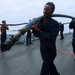 Naval personnel in dark blue coveralls are working on the deck of a military vessel at sea, carrying and handling a large blue hose with a metal nozzle/coupling attachment. The scene appears to be a training exercise or operational drill conducted on an overcast day. Multiple crew members are visible on the ship's deck, with the ocean visible in the background.