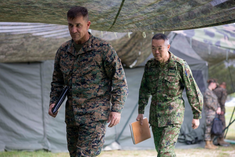 Two military officers in different camouflage uniforms walk together beneath a camouflage netting canopy near military field tents. The officer on the left wears U.S. Marines MARPAT digital camouflage with visible rank insignia and a name tape, while the officer on the right wears a different pattern consistent with Japan Ground Self-Defense Force (JGSDF) uniform. Both carry documents or folders, suggesting a joint military exercise or command briefing.