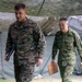 Two military officers in different camouflage uniforms walk together beneath a camouflage netting canopy near military field tents. The officer on the left wears U.S. Marines MARPAT digital camouflage with visible rank insignia and a name tape, while the officer on the right wears a different pattern consistent with Japan Ground Self-Defense Force (JGSDF) uniform. Both carry documents or folders, suggesting a joint military exercise or command briefing.
