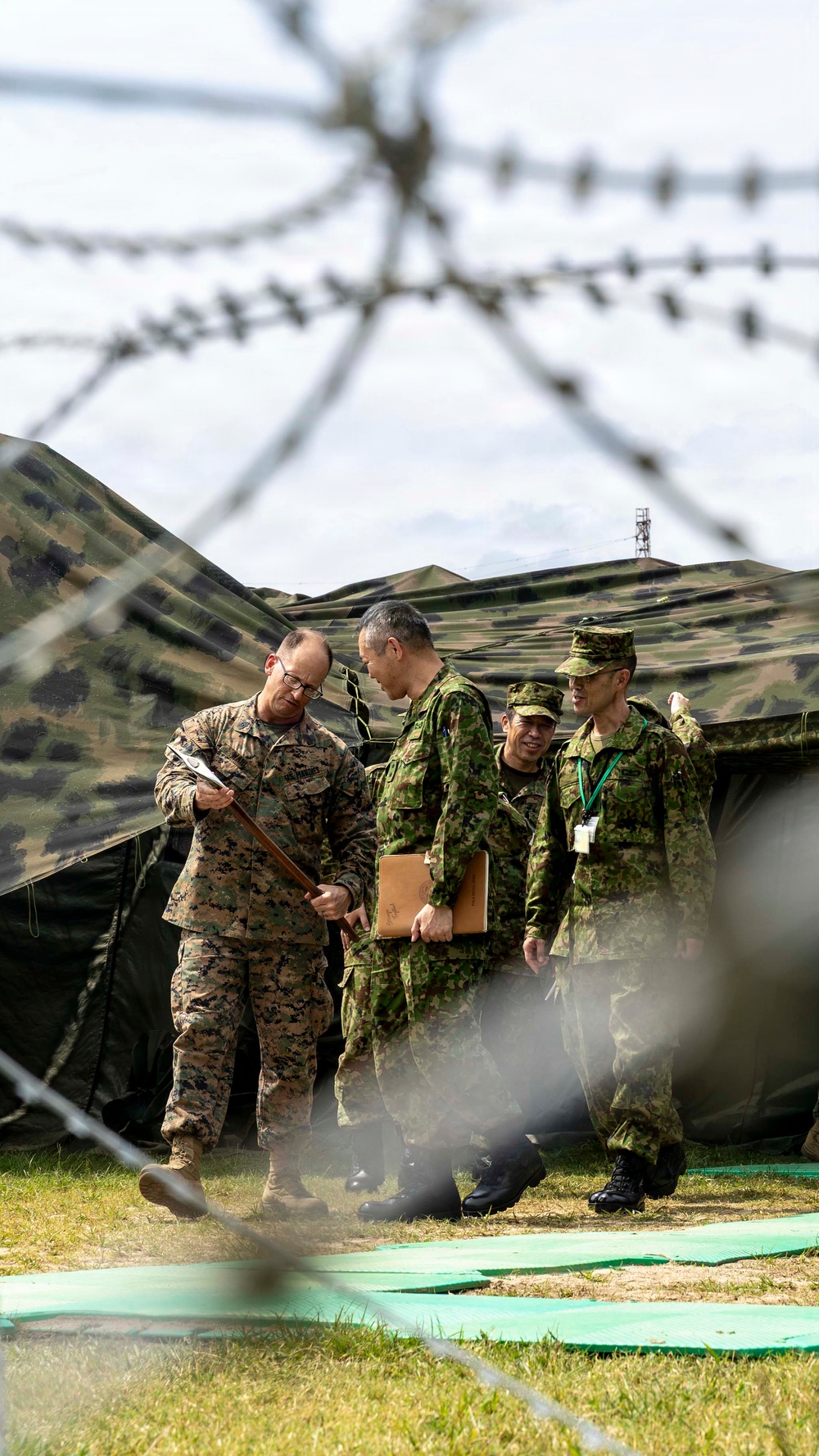 The image shows a group of military personnel from two different armed forces engaged in what appears to be a briefing or discussion near a camouflaged field tent. One soldier wearing MARPAT (US Marine Corps) digital camouflage is interacting with three soldiers wearing a different green camouflage pattern consistent with Japan Ground Self-Defense Force uniforms. The photo is taken through barbed wire in the foreground, which is intentionally blurred, suggesting a joint military exercise or bilateral training event.