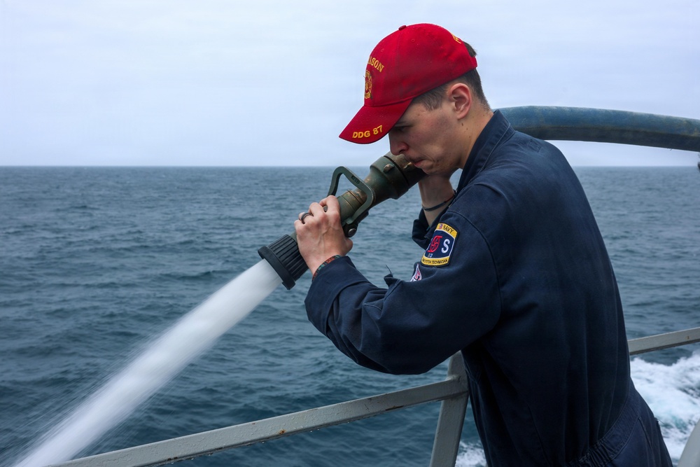 A U.S. Navy sailor aboard USS Mason (DDG 87) is operating a fire hose on the ship's deck, directing a high-pressure water stream over the side. The sailor is wearing Navy working uniform (NWU) coveralls and a red ball cap with 'Mason DDG 87' text. The ship appears to be underway at sea, visible from the wake in the background.