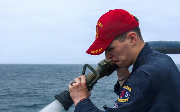 A U.S. Navy sailor aboard USS Mason (DDG 87) is operating a fire hose on the ship's deck, directing a high-pressure water stream over the side. The sailor is wearing Navy working uniform (NWU) coveralls and a red ball cap with 'Mason DDG 87' text. The ship appears to be underway at sea, visible from the wake in the background.