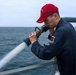 A U.S. Navy sailor aboard USS Mason (DDG 87) is operating a fire hose on the ship's deck, directing a high-pressure water stream over the side. The sailor is wearing Navy working uniform (NWU) coveralls and a red ball cap with 'Mason DDG 87' text. The ship appears to be underway at sea, visible from the wake in the background.