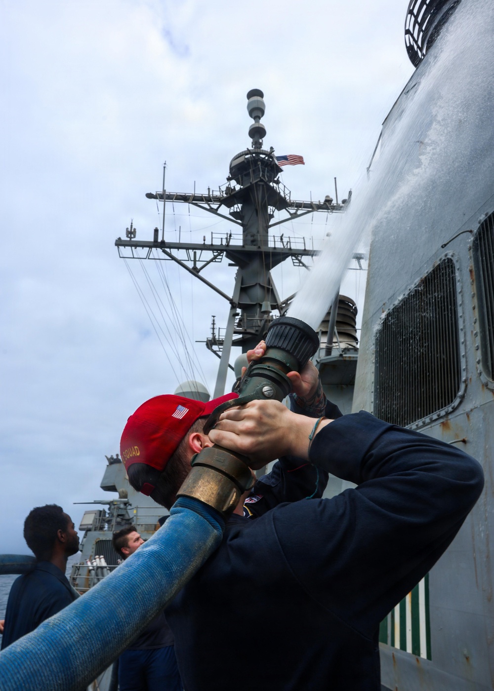 A sailor wearing a red cap with an American flag patch and a navy blue sweatshirt is operating a fire hose nozzle on the deck of a U.S. Navy warship. Water or foam is being discharged upward along the ship's superstructure, suggesting a damage control or firefighting drill. Additional crew members are visible in the background near the ship's mast, which displays an American flag.