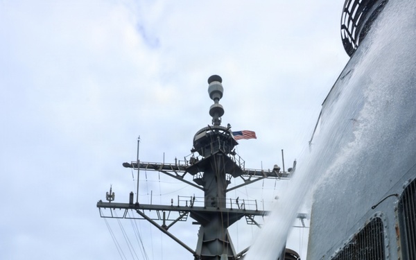 A sailor wearing a red cap with an American flag patch and a navy blue sweatshirt is operating a fire hose nozzle on the deck of a U.S. Navy warship. Water or foam is being discharged upward along the ship's superstructure, suggesting a damage control or firefighting drill. Additional crew members are visible in the background near the ship's mast, which displays an American flag.