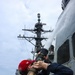 A sailor wearing a red cap with an American flag patch and a navy blue sweatshirt is operating a fire hose nozzle on the deck of a U.S. Navy warship. Water or foam is being discharged upward along the ship's superstructure, suggesting a damage control or firefighting drill. Additional crew members are visible in the background near the ship's mast, which displays an American flag.