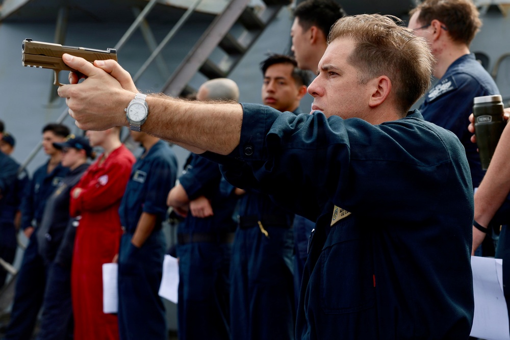 A military or naval personnel member in dark blue coveralls is aiming a semi-automatic pistol in a training or demonstration exercise on what appears to be the deck of a naval vessel. A group of similarly uniformed personnel stand in a line behind him, observing the activity. The setting and uniforms suggest a U.S. Navy or Coast Guard firearms qualification or safety training exercise.