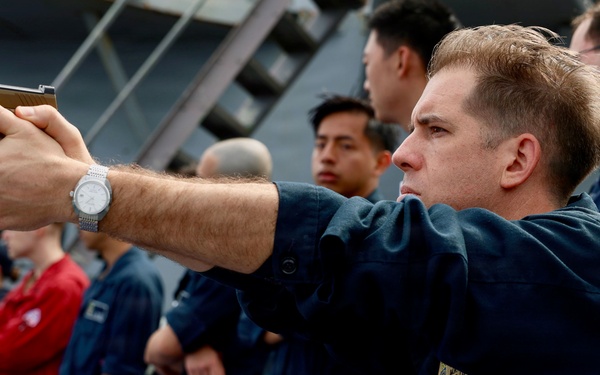A military or naval personnel member in dark blue coveralls is aiming a semi-automatic pistol in a training or demonstration exercise on what appears to be the deck of a naval vessel. A group of similarly uniformed personnel stand in a line behind him, observing the activity. The setting and uniforms suggest a U.S. Navy or Coast Guard firearms qualification or safety training exercise.